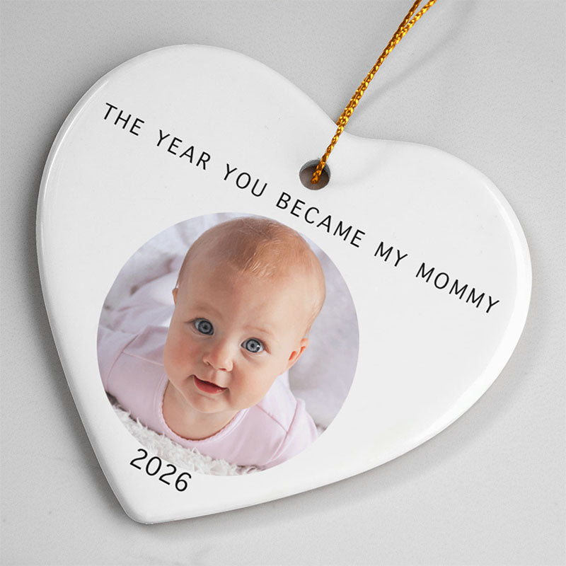 gift for new mom: a white heart-shaped ceramic ornament hanging from a gold string, printed with the text 'THE YEAR YOU BECAME MY MOMMY' and '2026', with a circular photo of a baby on the front.