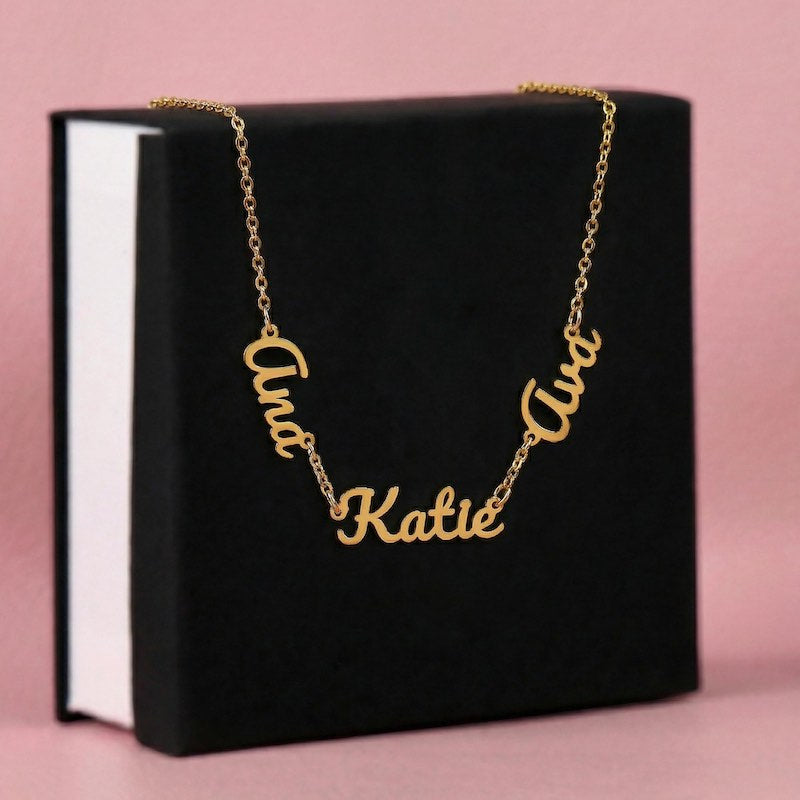 Gold name necklace with 'Ana''Katie'and 'Ava' cursive name pendants on a black box with pink background