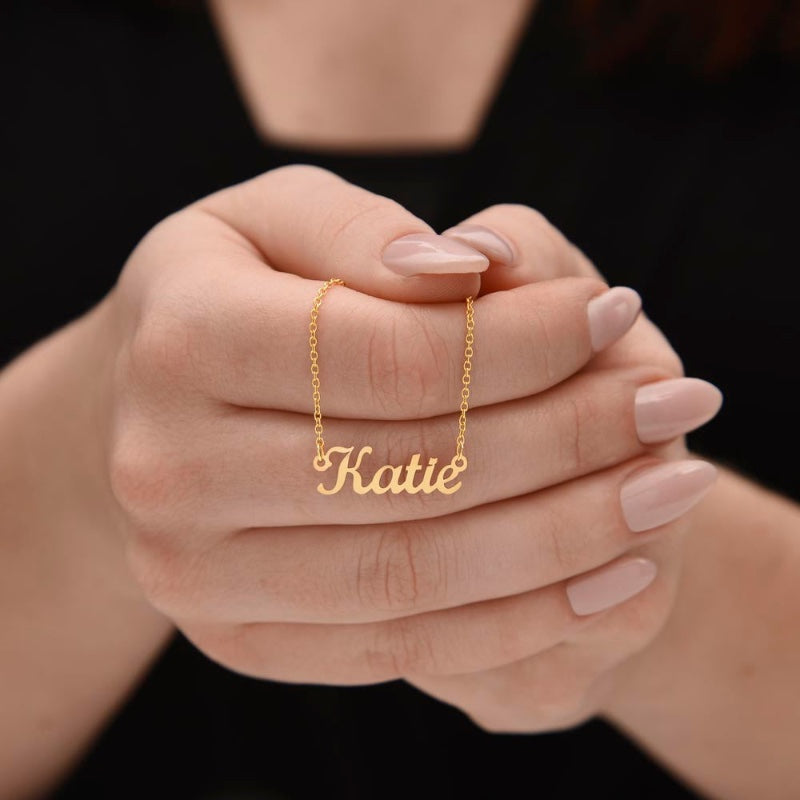 A close-up of a person's hands wearing a delicate gold chain necklace with a personalized nameplate that reads 'Katie' in cursive script.