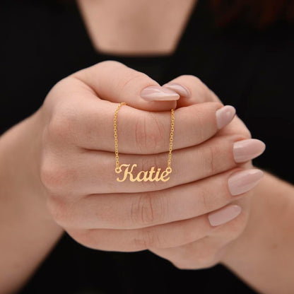 A close-up of a person's hands wearing a delicate gold chain necklace with a personalized nameplate that reads 'Katie' in cursive script.