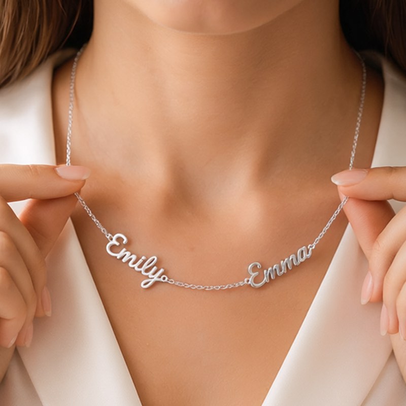 A close-up view of a person wearing a delicate, custom-made silver name necklace with 'Emily' and 'Emma' held by two hands. 