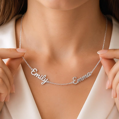 A close-up view of a person wearing a delicate, custom-made silver name necklace with 'Emily' and 'Emma' held by two hands. 