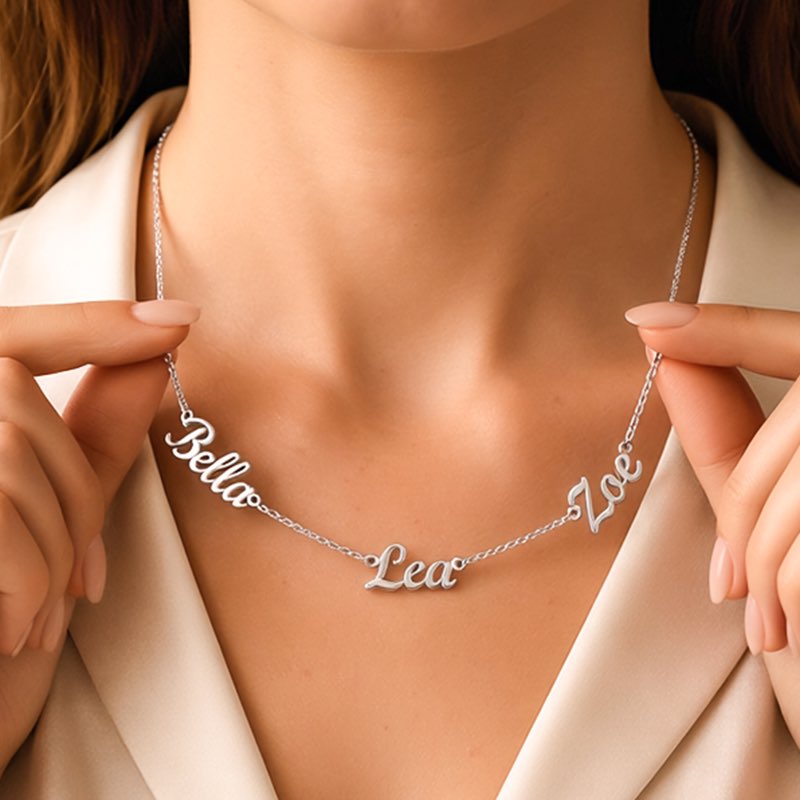 A close-up view of a person wearing a delicate, custom-made silver name necklace with cursive name pendants 'Bella', 'Lea', and 'Zoe' held by two hands.
