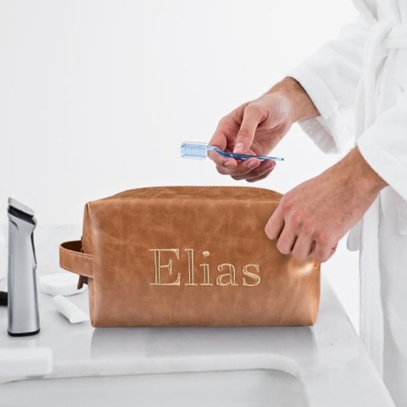 Brown leather travel toiletry bag embroidered with "Elias," shown in a bathroom setting with a person in a bathrobe holding a toothbrush.
