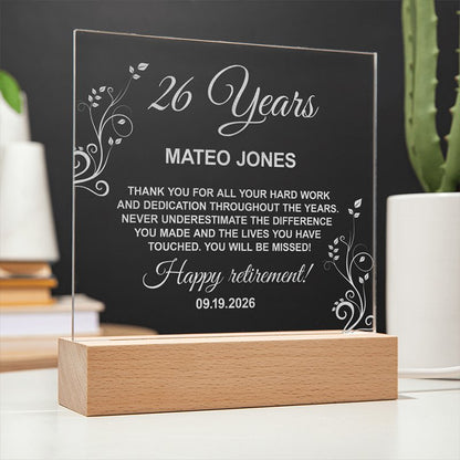 Custom clear acrylic retirement plaque mounted on a wooden base with white engraved floral accent designs, celebrating 26 years of dedication for Mateo Jones.