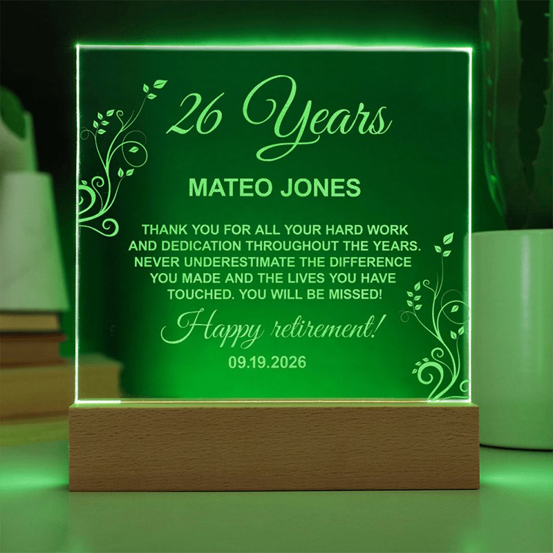 Illuminated personalized acrylic retirement plaque glowing green on a wooden LED base, featuring floral line engravings and a message honoring Mateo Jones for 26 years.