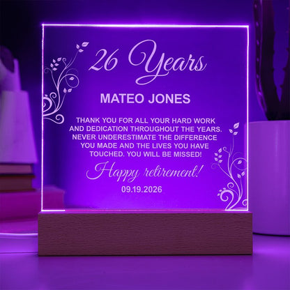 Illuminated personalized acrylic retirement plaque glowing purple on a wooden LED base, featuring floral line engravings and a message honoring Mateo Jones for 26 years.