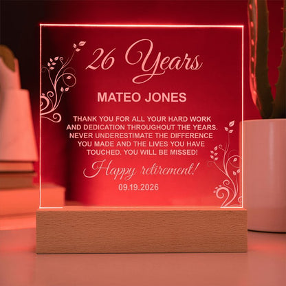 Illuminated personalized acrylic retirement plaque glowing red on a wooden LED base, featuring engraved floral accents and a message for Mateo Jones celebrating 26 years.