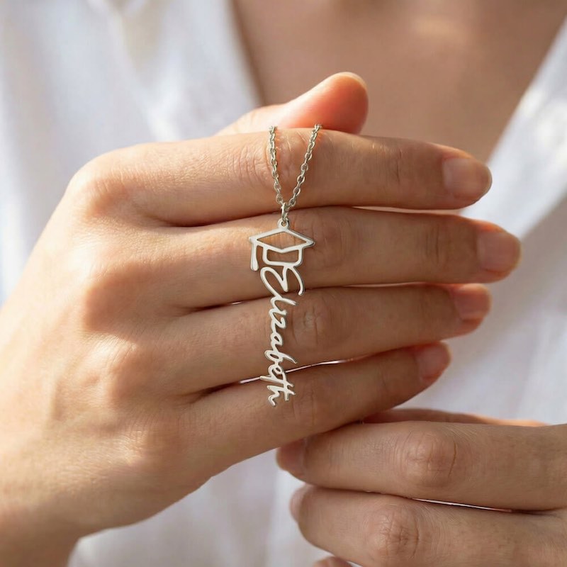 Silver graduation cap and name necklace held in a hand against a white background