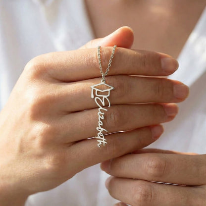 Silver graduation cap and name necklace held in a hand against a white background