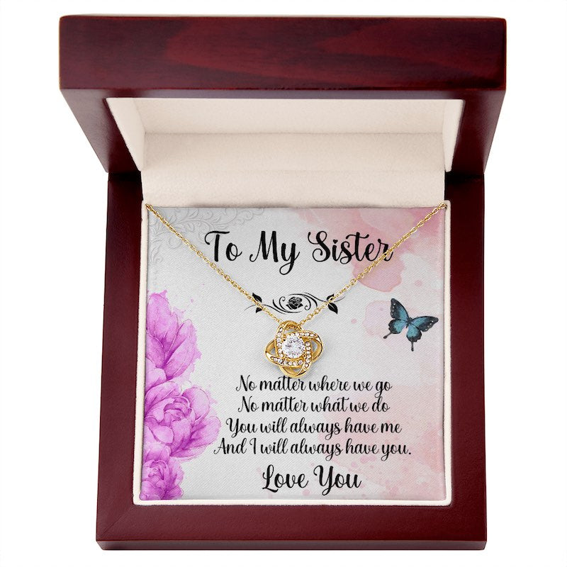 Gold love knot necklace displayed in an open luxury mahogany-style gift box with a sister message card featuring purple flowers and butterflies.