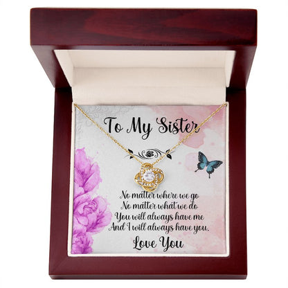 Gold love knot necklace displayed in an open luxury mahogany-style gift box with a sister message card featuring purple flowers and butterflies.