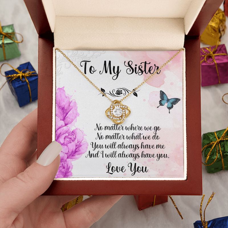 A hand holds an open luxury mahogany-style gift box containing a gold love knot necklace and a heartfelt sister message card, surrounded by miniature gift boxes.