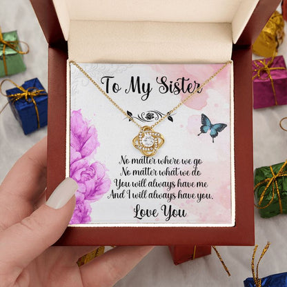 A hand holds an open luxury mahogany-style gift box containing a gold love knot necklace and a heartfelt sister message card, surrounded by miniature gift boxes.