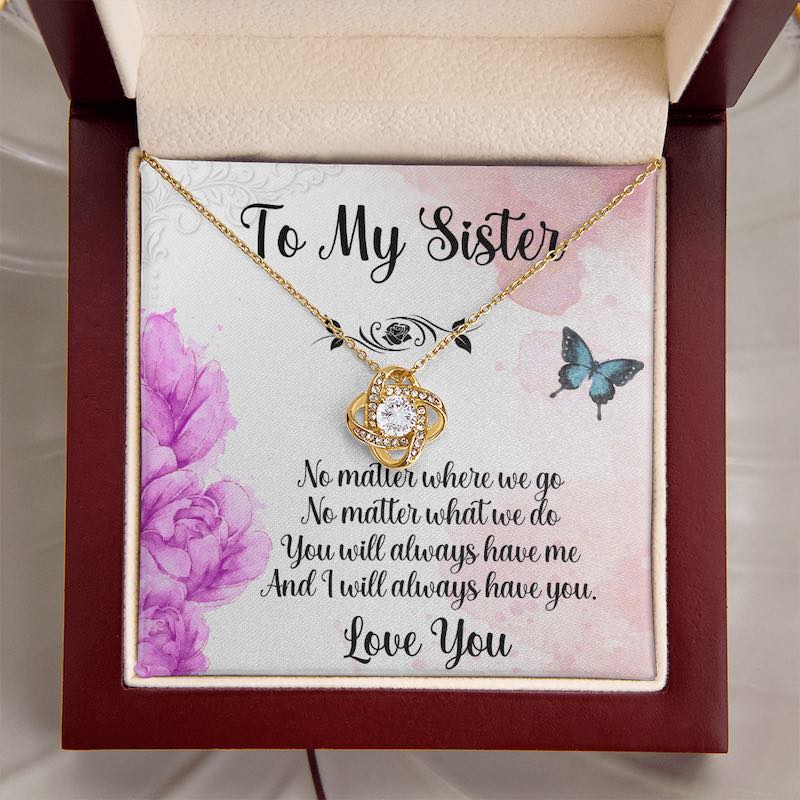 An elegant mahogany-style gift box set against a soft white silk background, showcasing a gold love knot necklace beside a heartfelt sister message card.