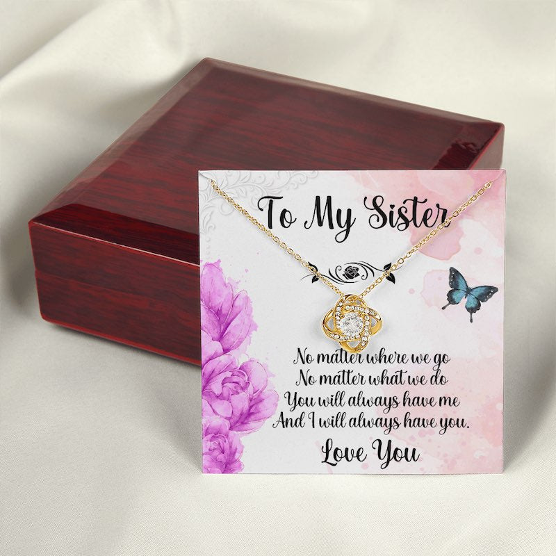 A closed luxury mahogany-style gift box positioned behind a gold love knot necklace displayed on a sister message card adorned with purple flowers and delicate butterflies.
