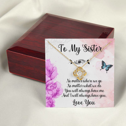A closed luxury mahogany-style gift box positioned behind a gold love knot necklace displayed on a sister message card adorned with purple flowers and delicate butterflies.