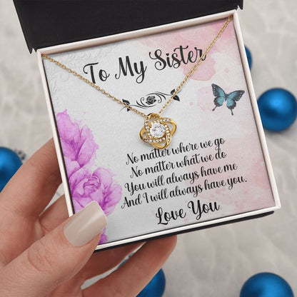 Hand holding an open white gift box displaying a gold love knot necklace with a sister message card featuring purple flowers and butterflies.   
