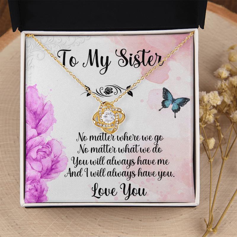 Gifts for sister: White gift box resting on a wood slice near dried flowers, displaying a gold love knot necklace with a sister message card featuring purple flowers and butterflies.