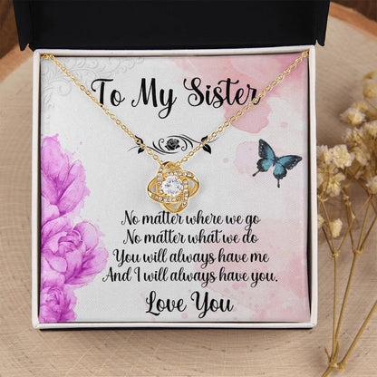 Gifts for sister: White gift box resting on a wood slice near dried flowers, displaying a gold love knot necklace with a sister message card featuring purple flowers and butterflies.