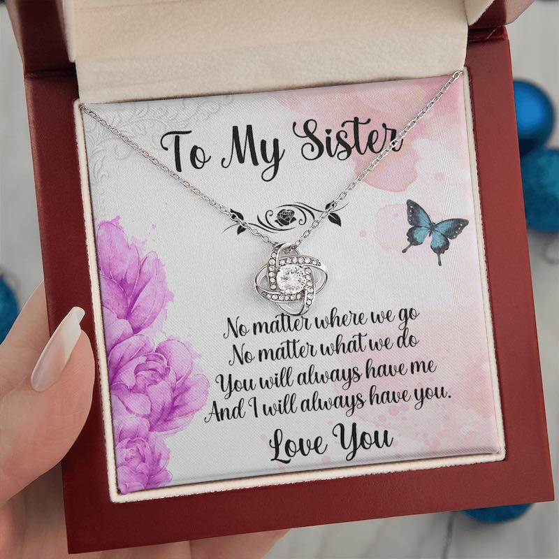 Hand holding an open luxury mahogany-style gift box to present a silver love knot necklace with a sister message card featuring purple flowers and butterflies.