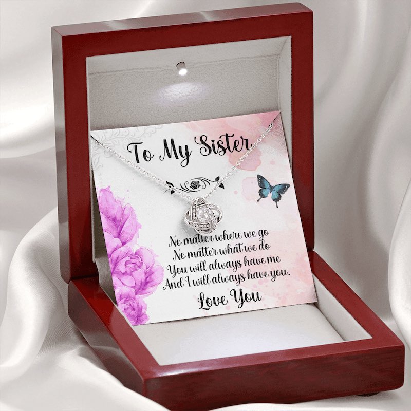 Personalized sister gifts: Silver love knot necklace displayed in an illuminated luxury mahogany-style gift box resting on white silk, with a sister message card featuring purple flowers and butterflies.