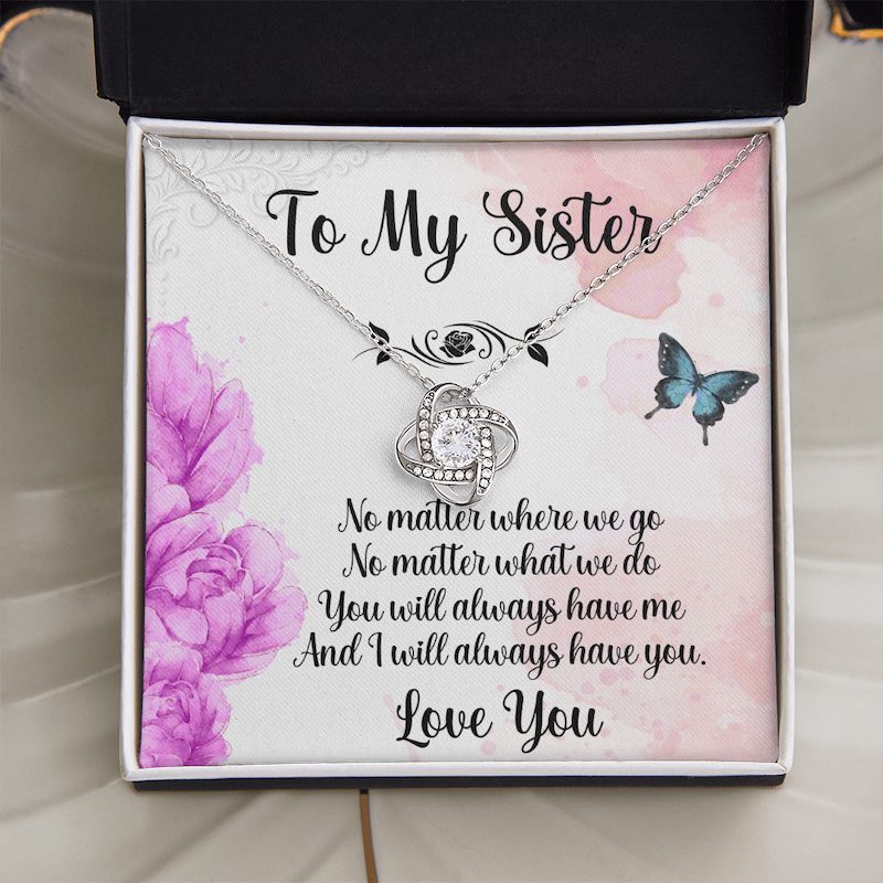 An open white gift box resting on a white surface, displaying a silver love knot necklace with a sister message card.