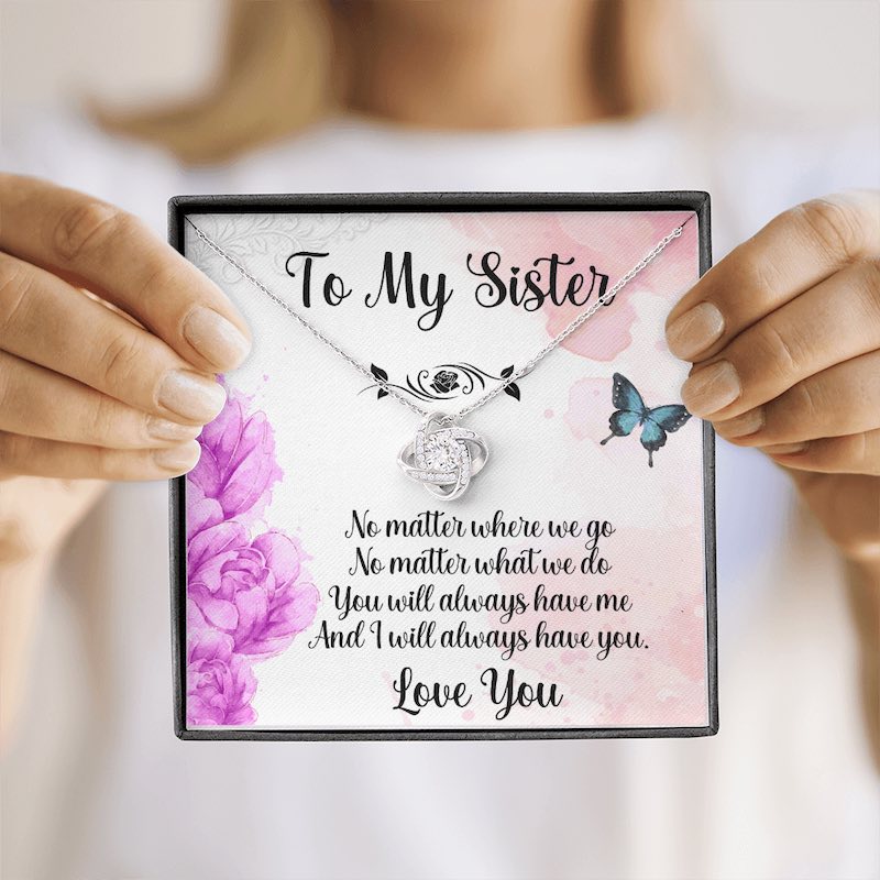 A person holds a white gift box against a softly blurred background, showcasing a silver love knot necklace alongside a heartfelt sister message card.