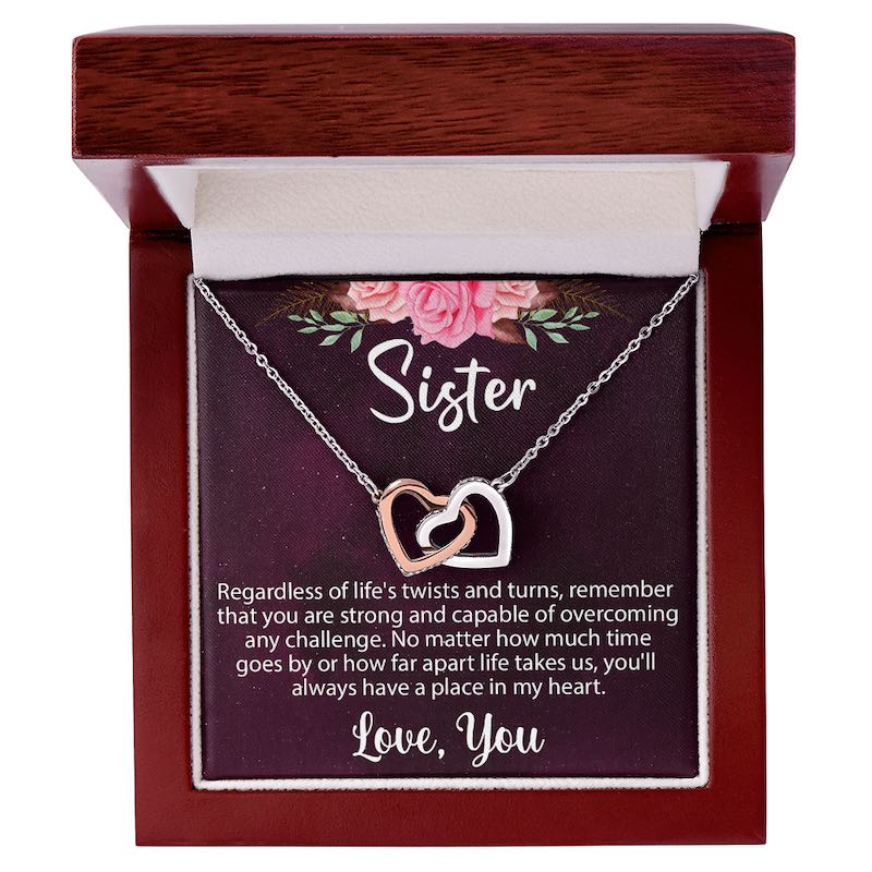 An open luxury mahogany-style wooden gift box displaying a silver and rose-gold interlocking heart necklace on a sentimental "Sister" message card.