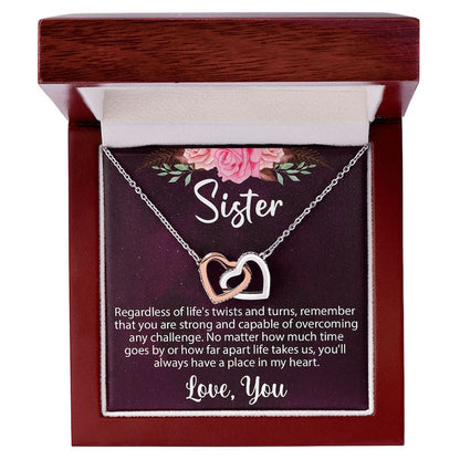 An open luxury mahogany-style wooden gift box displaying a silver and rose-gold interlocking heart necklace on a sentimental "Sister" message card.