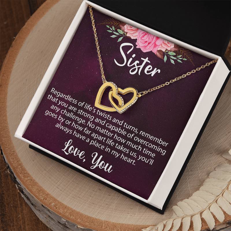 A gold interlocking heart necklace inside an open gift box with a sentimental "Sister" message card, displayed on a rustic wood slice with dried botanical accents.