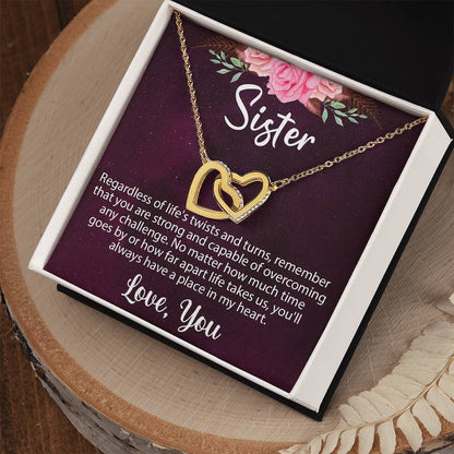 A gold interlocking heart necklace inside an open gift box with a sentimental "Sister" message card, displayed on a rustic wood slice with dried botanical accents.
