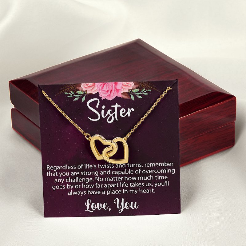A gold interlocking heart necklace on a sentimental "Sister" message card displayed in front of a closed luxury mahogany-style wooden gift box on a white satin background