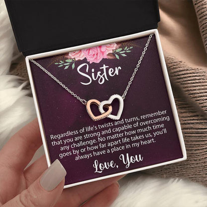 A hand holding an open gift box showcasing a rose gold and silver interlocking heart necklace on a sentimental "Sister" message card against a cozy knit background.