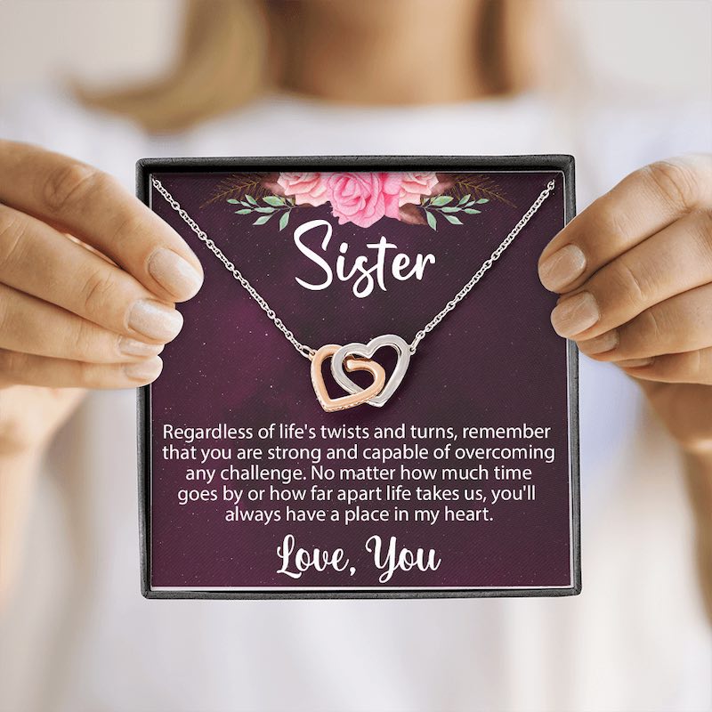 A person holding an open gift box displaying a rose-gold and silver interlocking heart necklace on a sentimental "Sister" message card.