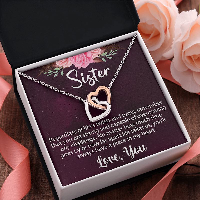 An open gift box displaying a rose gold and silver interlocking heart necklace on a sentimental "Sister" message card, styled with soft pink roses and a ribbon on a rustic wooden surface.