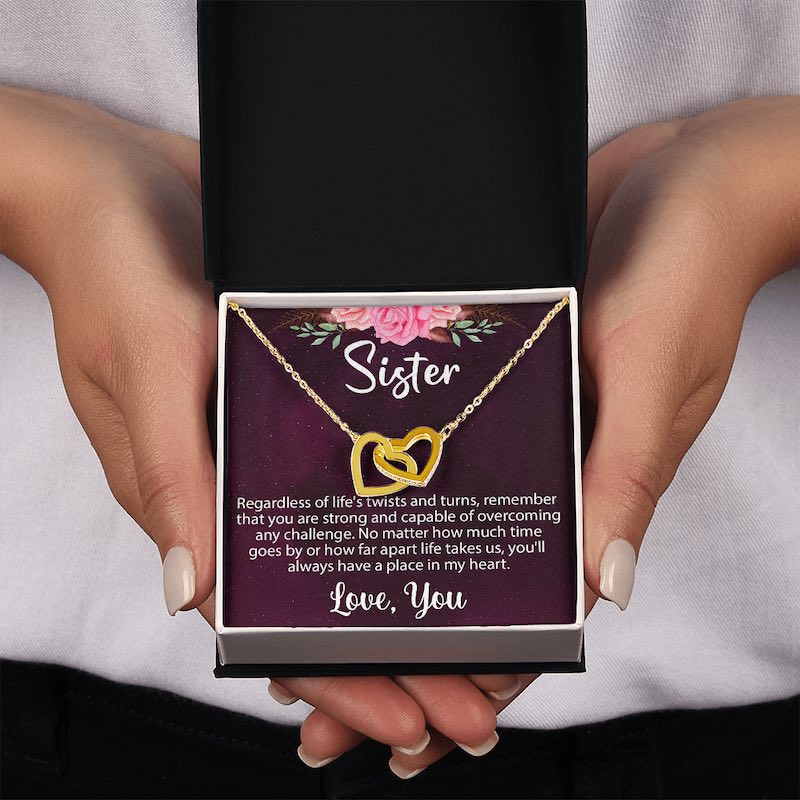 Hands holding an open gift box displaying a gold interlocking heart necklace resting on a sentimental "Sister" message card.