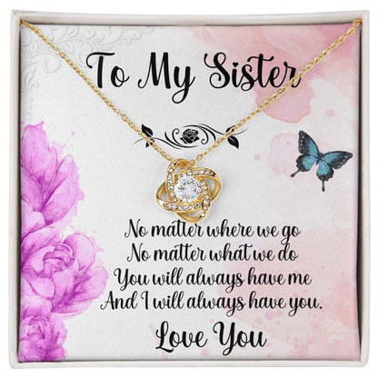 Open white gift box displaying a gold love knot necklace with a purple floral and butterfly sister message card.   