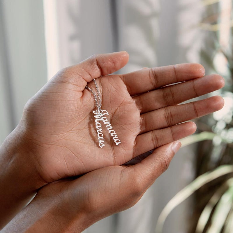 two hands keeping a name necklace silver featuring two vertical name pendants on a blurry light green background.