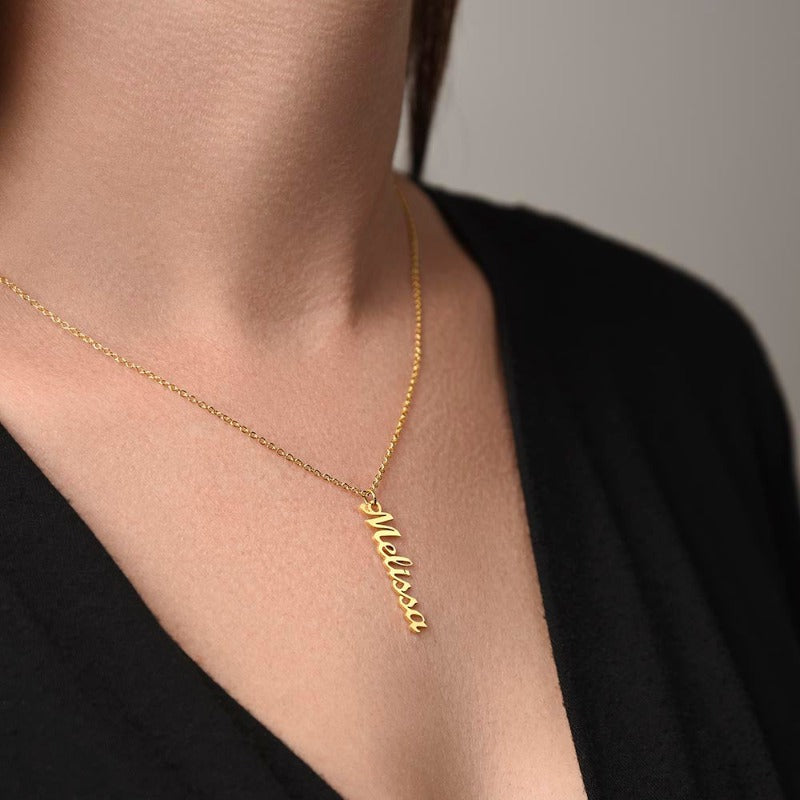 A gold name necklace with a vertical pendant displayed on a woman's chest.