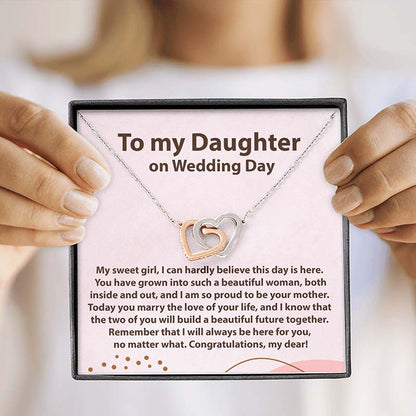 A silver double-heart necklace with one rose-gold heart and one silver heart is displayed on a pink gift card that reads 'To my Daughter on Wedding Day.' The card is held by hands with manicured nails against a softly blurred light background.
