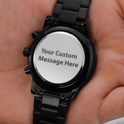 A close-up of a black metal watch held in a hand, showing the back of the case with a silver circular insert that reads 'Your Custom Message Here'.
