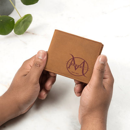 Personalized brown leather men's bifold wallet engraved on the bottom right corner with a circular monogram featuring a large letter 'M', the name "MORGAN", and "EST. 2024", held by hands, on a light marble table.