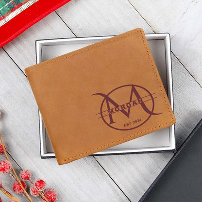 Brown leather men's wallet custom engraved with a circular monogram design featuring a large 'M', "MORGAN", and "EST. 2024", resting inside a white box lid on a white wooden background with festive red berries.   