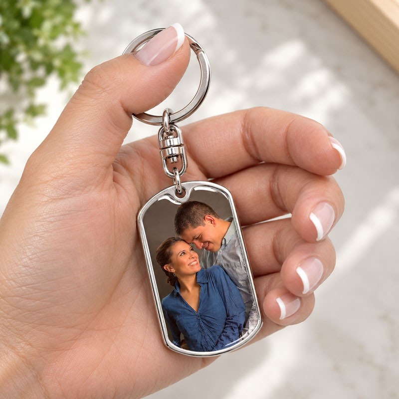A hand holds a silver metal keychain featuring a custom rectangular photo insert of a couple, set against a softly blurred light background.